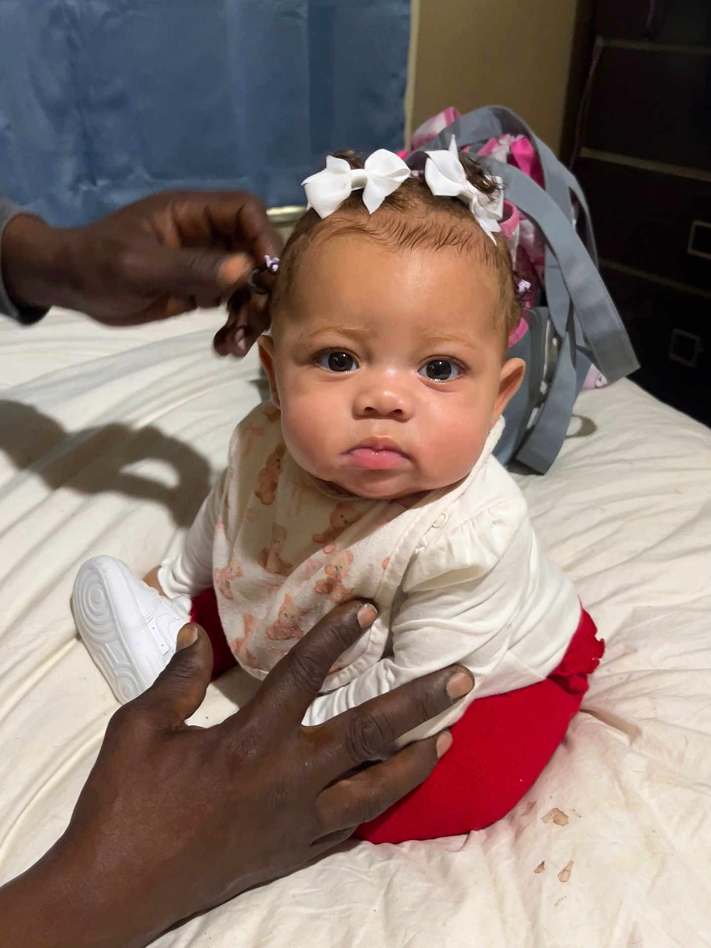 Amari is registered to the contest to win money with this photo: baby, child, hands, bed, white_bows, red_pants, bib, backpack, indoor, person, face, eyes, clothing, sitting, skin, hair, expression, adult_hands, soft_lighting, curly_hair
