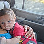 baby, child, infant, blue_eyes, red_onesie, cartoon_print, adult_hand, car_interior, window, seat, tattoo, skin, comfort, resting, close_up, portrait, natural_light, caring, family, tender_moment