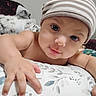baby, child, striped_hat, hand, pillow, blanket, face, eyes, indoor, closeup, infant, curious, cute, person, soft_lighting, skin, expression, cozy, bedroom, comfort