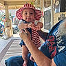 Emiliano is registered to the contest to win money with this photo: baby, child, infant, hat, striped_hat, american_flag, patriotic, tattooed_hand, beard, adult, porch, outdoor, decorations, clothing, blue_shirt, smiling, person, holding, foot, face