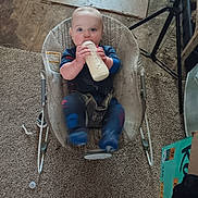 Jesus is registered to the contest to win money with this photo: baby, infant, child, bottle, milk, feeding, bouncer, seated, blue_clothing, wide_eyes, portrait, indoor, living_room, carpet, tripod, furniture, household, floor, smiling, candid
