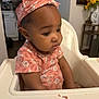 baby, child, curly_hair, expression, floral_pattern, flowers, furniture, headband, high_chair, home, indoor, kitchen, person, pink_clothing, portrait, sitting, table, toddler, vase, yellow_flowers