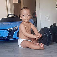 Benett participe au concours pour gagner de l'argent avec cette photo : toddler, baby, diaper, indoor, floor, toy_car, blue_car, dumbbell, weight, exercise_equipment, child, sitting, bare_chest, home, room, wooden_floor, serious_expression, fitness, play, young_child