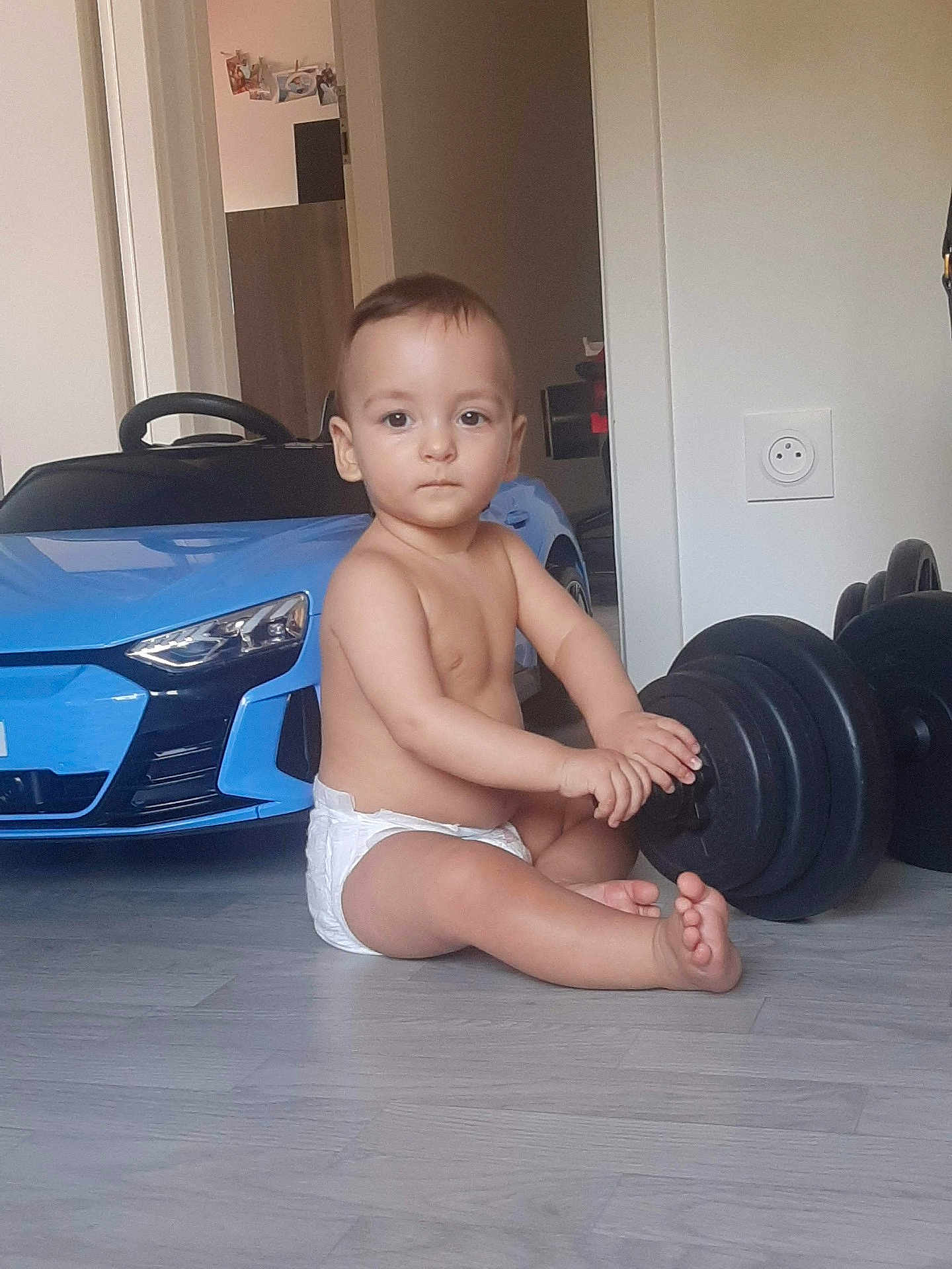 Benett participe au concours pour gagner de l'argent avec cette photo : toddler, baby, diaper, indoor, floor, toy_car, blue_car, dumbbell, weight, exercise_equipment, child, sitting, bare_chest, home, room, wooden_floor, serious_expression, fitness, play, young_child