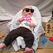 Eric is registered to the contest to win money with this photo: baby, sunglasses, toy_cars, shirt, towel, blanket, car, floor, sign, cardboard, person, child, clothing, footwear, indoor, relaxing, cute, playing, funny, casual