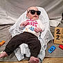baby, sunglasses, toy_cars, shirt, towel, blanket, car, floor, sign, cardboard, person, child, clothing, footwear, indoor, relaxing, cute, playing, funny, casual