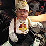 baby, child, thanksgiving, hat, mittens, cooking_pot, stove, kitchen, cute, infant, sitting, clothing, holiday, tableware, appliance, person, indoor, face, expression, warm