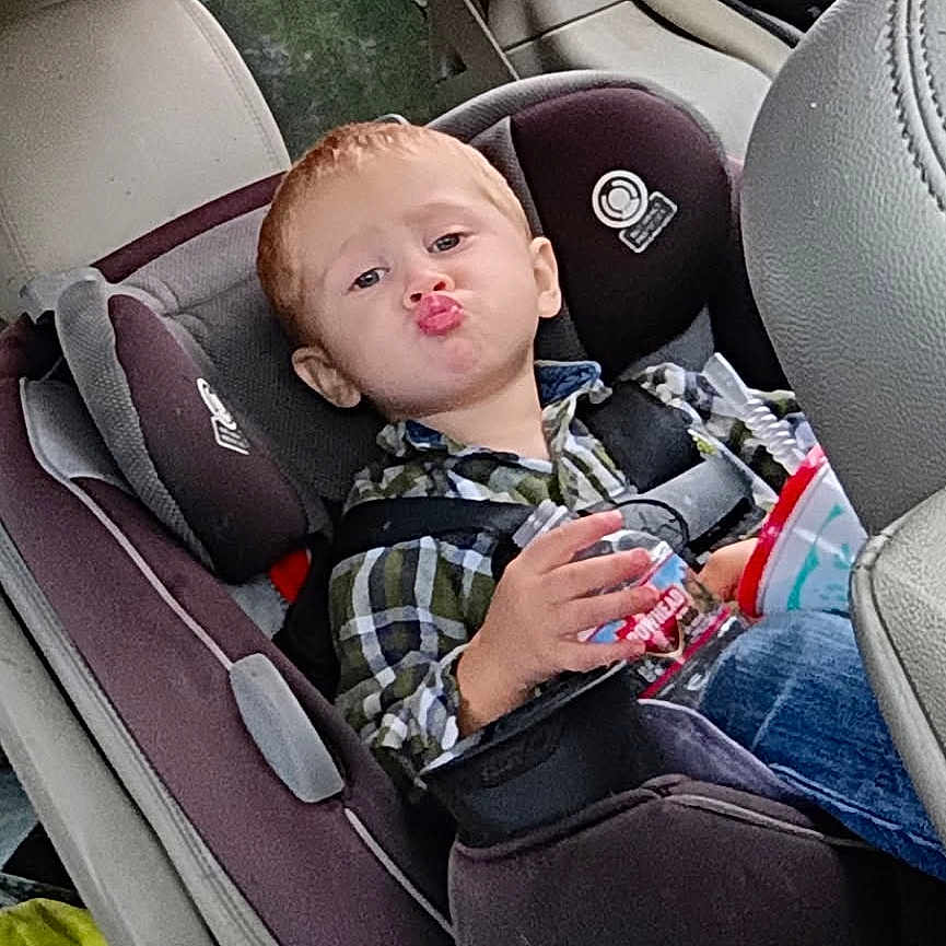 Dantyvious is registered to the contest to win money with this photo: blue_jeans, car_door, car_seat, car_seat_harness, child, child_safety, cup, funny_face, indoor, person, plaid_shirt, puckered_lips, red_hair, seat, seatbelt, toddler, toy, tree_outside, vehicle_interior, window