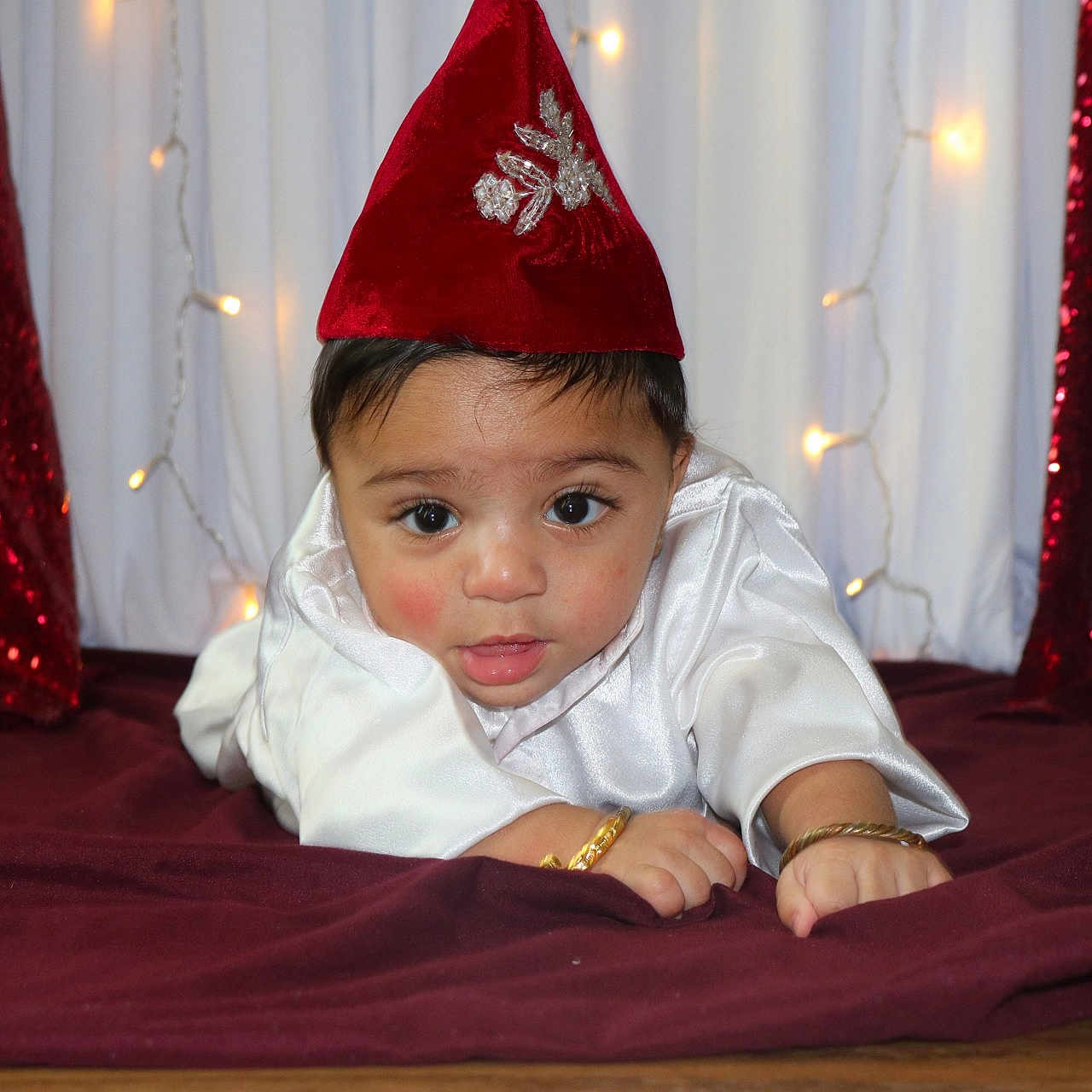 Arnish joined the competition — help win amazing prizes! baby, bracelet, child, closeup, curious_expression, cute, decor, fabric, face, festive, hat, holiday_lights, indoors, infant, lying_down, portrait, red_hat, sequins, sparkle, white_clothing
