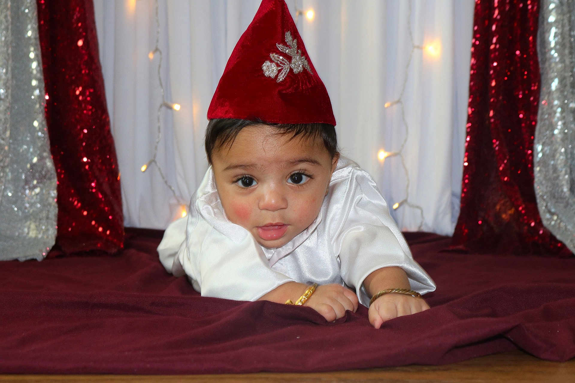 Arnish joined the competition — help win amazing prizes! baby, bracelet, child, closeup, curious_expression, cute, decor, fabric, face, festive, hat, holiday_lights, indoors, infant, lying_down, portrait, red_hat, sequins, sparkle, white_clothing