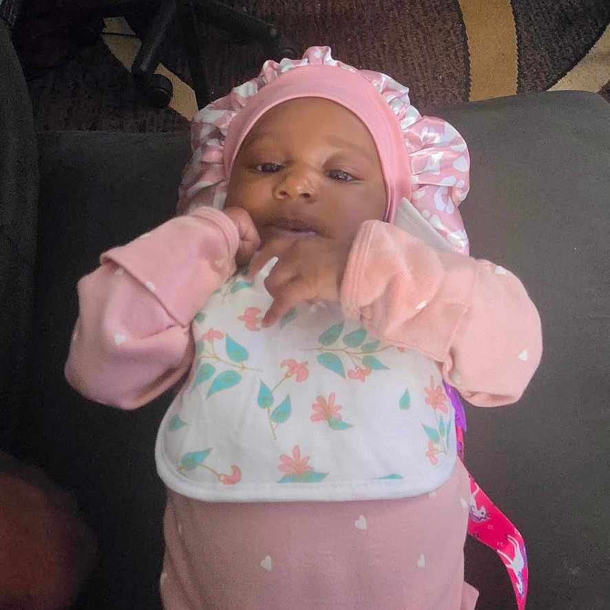 Niriyah Christina is registered to the contest to win money with this photo: baby, infant, child, portrait, pink_clothing, bonnet, pajamas, sitting, lying_down, smiling, face, hands, floral_pattern, couch, lap, indoor, office_chairs, carpet, cables, home_photo