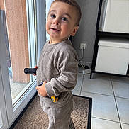 Noham a rejoint le concours — aidez-le/la à gagner de superbes lots ! toddler, child, toy_cars, indoor, window, glass_door, curious, standing, beige_outfit, ribbed_clothing, white_sneakers, hair_spiky, carpet, tile_floor, storage_boxes, playful, person, young_child, holding, smiling