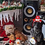 dog, christmas, holiday, plush_toys, santa_claus, reindeer_toy, gingerbread_man, penguin_toy, snowman_headband, speaker, decorations, blanket, festive, indoor, floor, cute, black_dog, holiday_decor, toy, winter