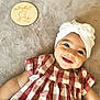 baby, smiling, headband, bow, dress, checkered, plaque, wooden, milestone, age, blanket, soft, lying_down, cute, happy, child, infant, portrait, indoor, closeup