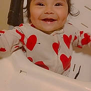 Luna is registered to the contest to win money with this photo: baby, cheerful, child, clothing, cute, expression, face, happy, heart_pattern, high_chair, indoor, infant, onesie, person, portrait, red, seat, smiling, white, young_child