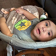 Julian is registered to the contest to win money with this photo: toddler, baby_rocker, blanket, indoor, child, curious_expression, clothing, face, person, seat_belt, wooden_floor, couch, soft_texture, comfort, casual_wear, resting, young_child, portrait, seat, relaxation