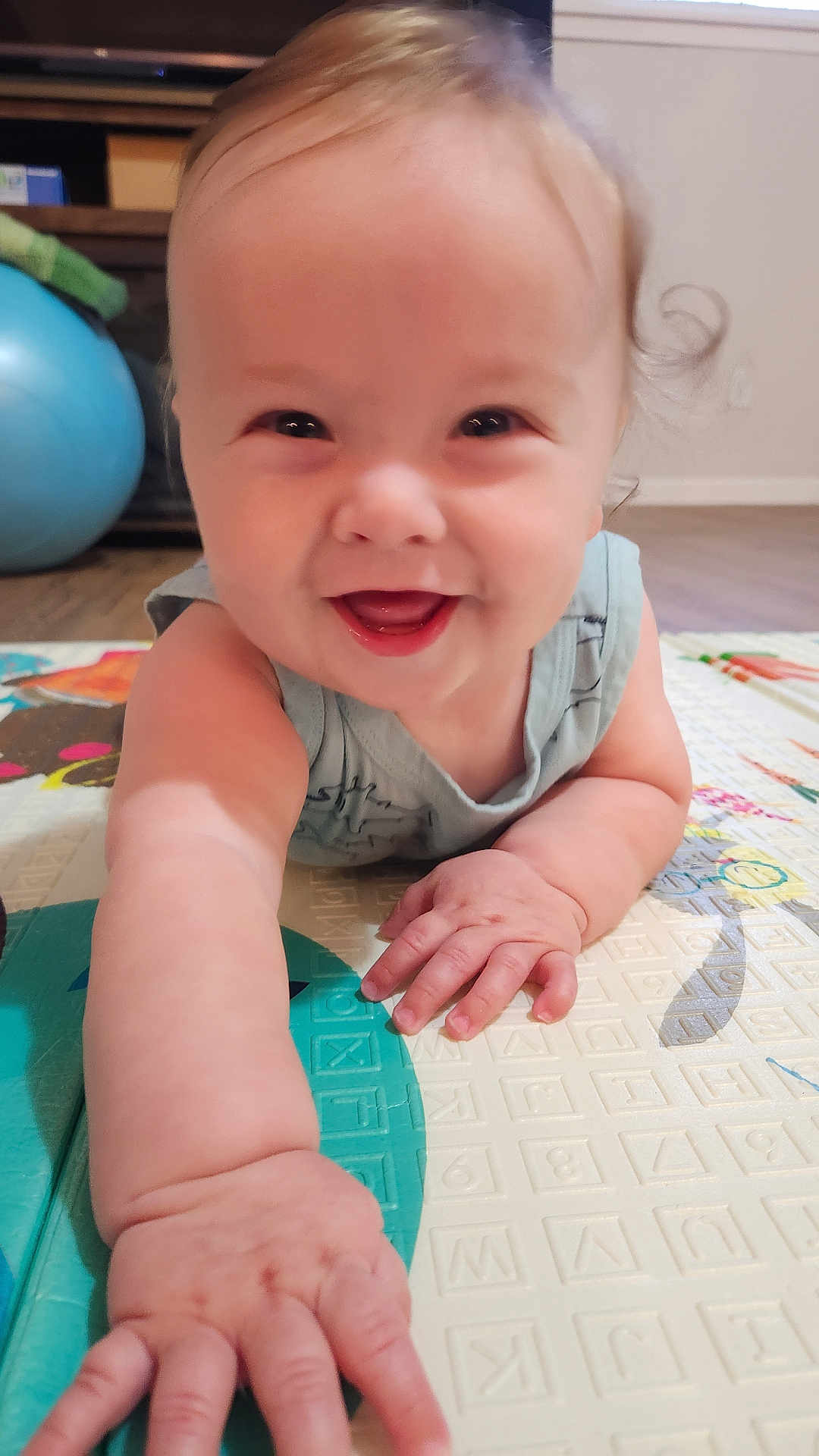 Finnegan is registered to the contest to win money with this photo: baby, smiling, crawling, child, play_mat, hand, face, indoor, floor, exercise_ball, happy, person, infant, skin, furniture, home, curly_hair, closeup, cute, young_child