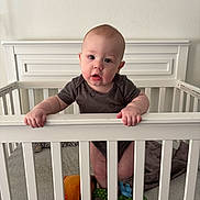 Beckham is registered to the contest to win money with this photo: baby, crib, infant, child, onesie, toy, blanket, bed, hand, face, person, indoor, white, wood, curious, standing, soft_toy, expression, grip, room