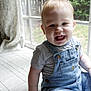 baby, child, smiling, denim_overalls, striped_shirt, window, natural_light, curtain, bench, indoor, happy, face, person, toddler, cushion, hair, skin, sitting, mouth, teeth