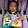 baby, child, car_seat, pink_bow, soft_book, panda, monkey, colorful, cute, infant, seatbelt, clothing, foot, hand, indoors, portrait, person, toy, fabric, sitting