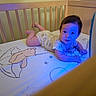 Anastasia is registered to the contest to win money with this photo: baby, crib, wooden_crib, infant, blue_light, cartoon_bear, sheet, cute, child, tummy_time, playful, indoors, furniture, bedroom, face, person, curious, young_child, smiling, pajamas