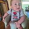 baby, child, infant, smiling, portrait, sitting, high_chair, gray_shirt, red_text, clothing, face, eyes, indoor, home, window, mirror, furniture, wood_floor, daylight, happy