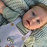baby, child, infant, portrait, face, eyes, smiling, lying_down, blanket, knitted_blanket, clothing, overalls, striped_clothing, green_shirt, tractor, embroidered_design, buttons, blond_hair, indoor, close_up