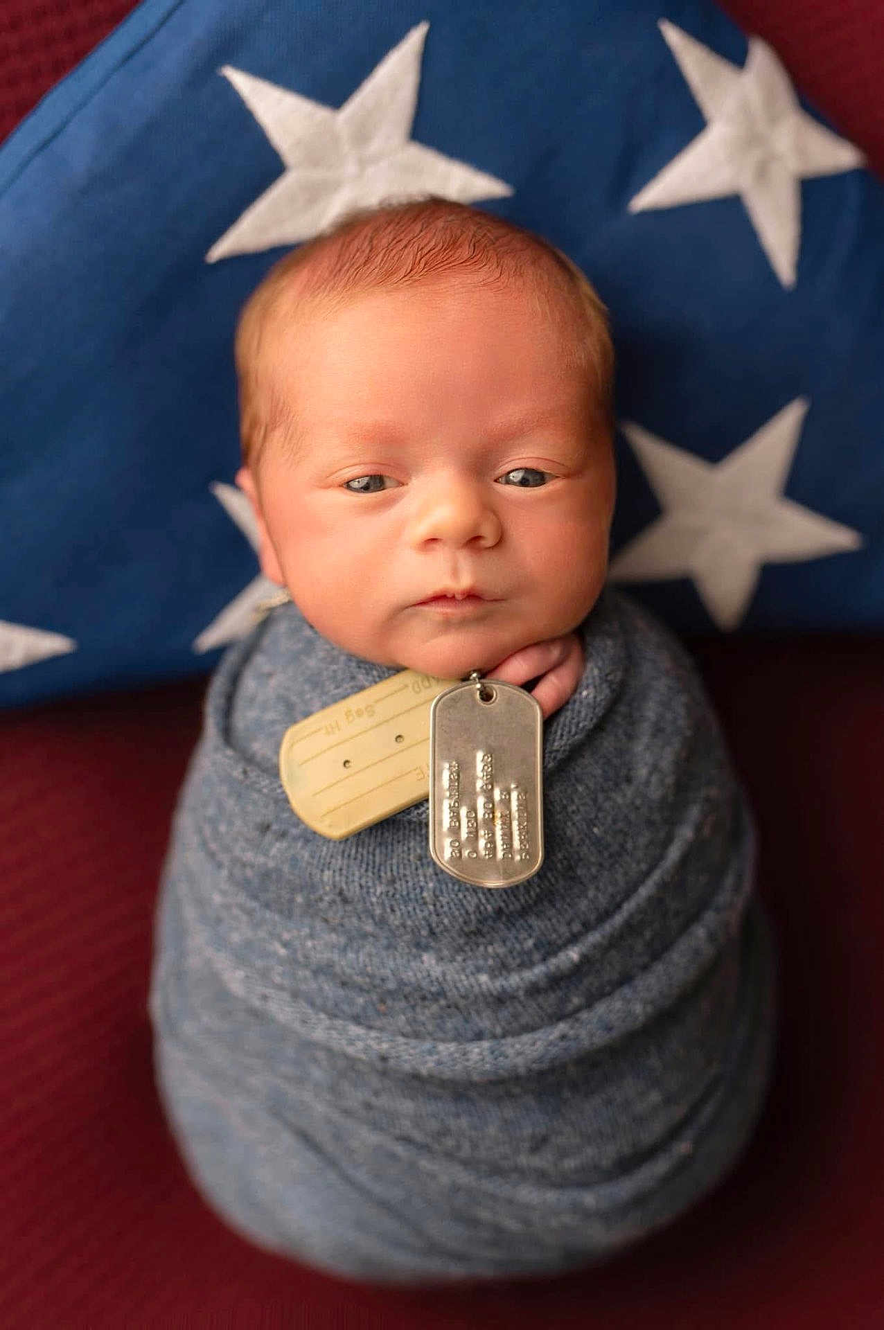 Danny joined the competition — help win amazing prizes! newborn, baby, swaddle, blue, dog_tags, portrait, face, infant, pillow, stars, knitted, wrapped, serious_expression, closeup, indoor, soft_light, red_background, cute, child, person
