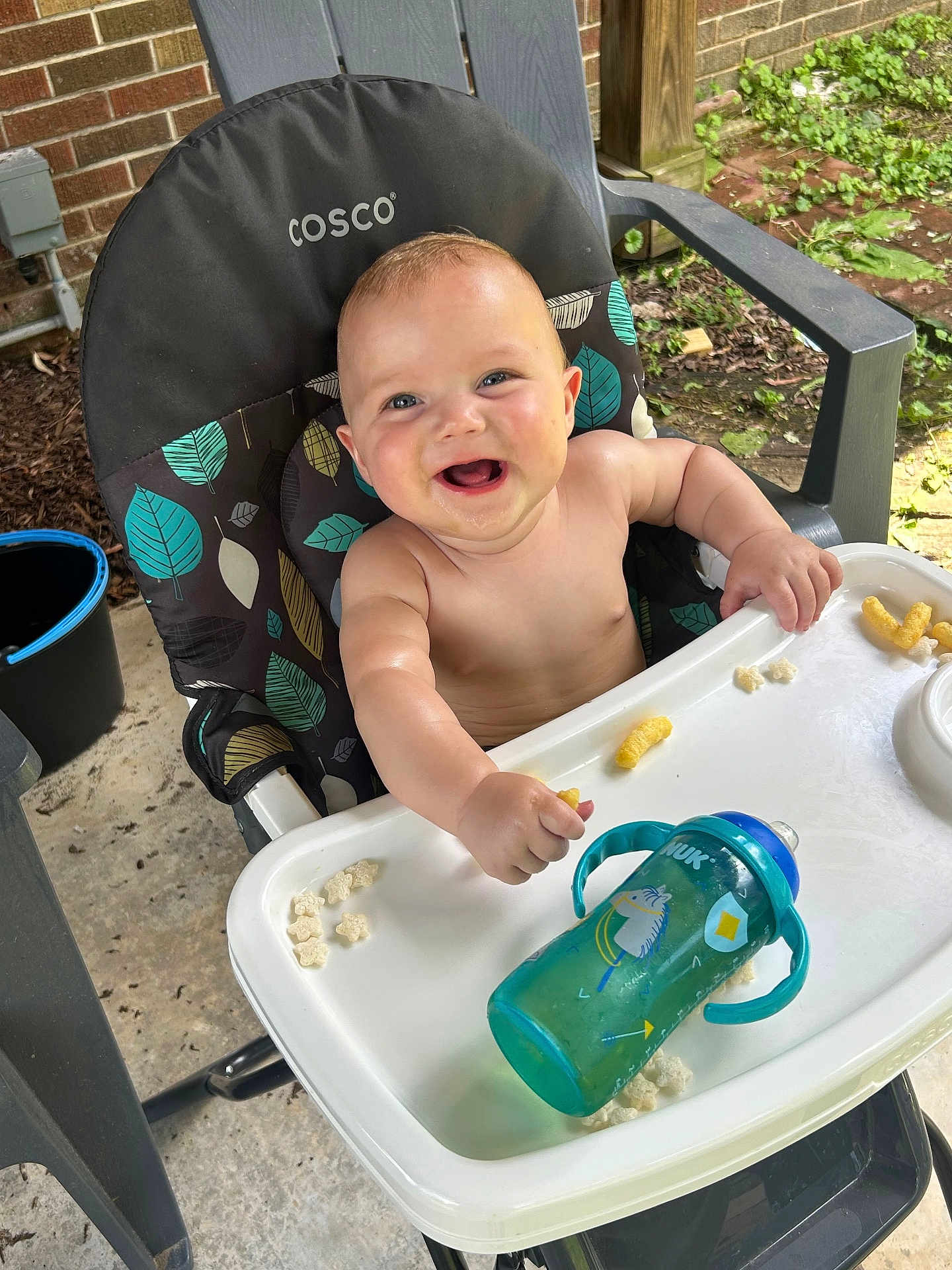 Kaelen joined the competition — help win amazing prizes! baby, high_chair, smiling, snacks, sippy_cup, outdoor, leaf_pattern, happy, child, food, plastic_chair, concrete_floor, brick_wall, greenery, person, infant, playful, feeding, toddler, sunlight
