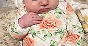 Willow is registered to the contest to win money with this photo: baby, infant, child, floral_clothing, onesie, cushion, blanket, wooden_headboard, indoor, curious, sitting, hand, face, person, patterned, colorful, strap, soft_lighting, cozy, portrait
