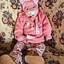 baby, boots, bow, cushion, eyes, face, floral_pattern, hands, headband, home_interior, infant, legs, mouth, pacifier, patterned_fabric, pink_outfit, portrait, sofa, upholstery, wood_paneling
