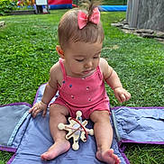 Mylee is registered to the contest to win money with this photo: baby, child, pink_bow, pink_romper, wooden_toy, grass, blue_mat, outdoor, backyard, inflatable_play_structure, banner, summer, playtime, cute, sitting, toddler, toy, daylight, nature, fun
