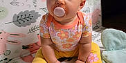 Mylee is registered to the contest to win money with this photo: baby, pacifier, child, sitting, play_mat, colorful, floral_clothing, yellow_pants, soft_shoes, curly_hair, indoors, cute, toy, patterned_mat, innocent_look, floor_seating, small_hands, bright_colors, young_child, casual_wear