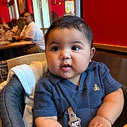 Mateo is registered to the contest to win money with this photo: baby, child, high_chair, blue_shirt, curious_eyes, chubby_cheeks, indoor, restaurant, wooden_table, people, background_blur, red_wall, short_hair, toddler, infant, cute, seated, skin, hand, clothing