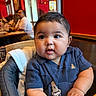 baby, child, high_chair, blue_shirt, curious_eyes, chubby_cheeks, indoor, restaurant, wooden_table, people, background_blur, red_wall, short_hair, toddler, infant, cute, seated, skin, hand, clothing