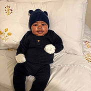 Theodore is registered to the contest to win money with this photo: baby, infant, bed, pillow, blanket, hat, mittens, clothing, cute, portrait, indoor, smiling, cozy, black_outfit, white_bedding, floral_embroidery, child, person, young, resting