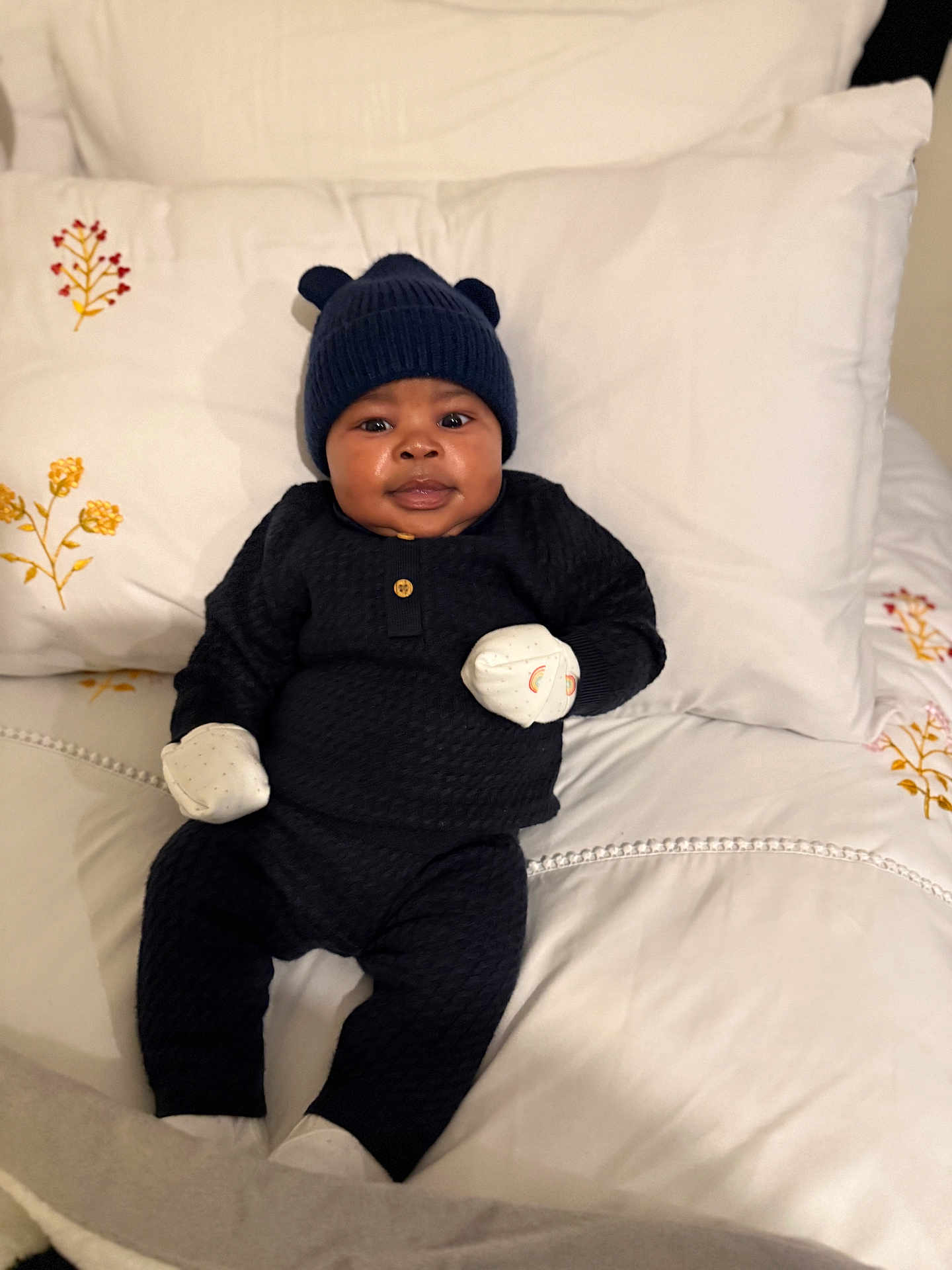 Theodore is registered to the contest to win money with this photo: baby, infant, bed, pillow, blanket, hat, mittens, clothing, cute, portrait, indoor, smiling, cozy, black_outfit, white_bedding, floral_embroidery, child, person, young, resting