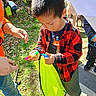 activity, adult, bag, boy, casual_clothing, checkered_shirt, child, curious, daylight, grass, leaves, marker, nature, outdoor, people, play, red_shirt, sneakers, sunlight, yellow