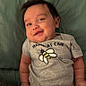 baby, infant, smiling, onesie, bee, text_on_clothing, chubby_cheeks, lying_down, green_blanket, cute, happy, child, portrait, skin, hand, head, hair, casual, indoors, soft_lighting