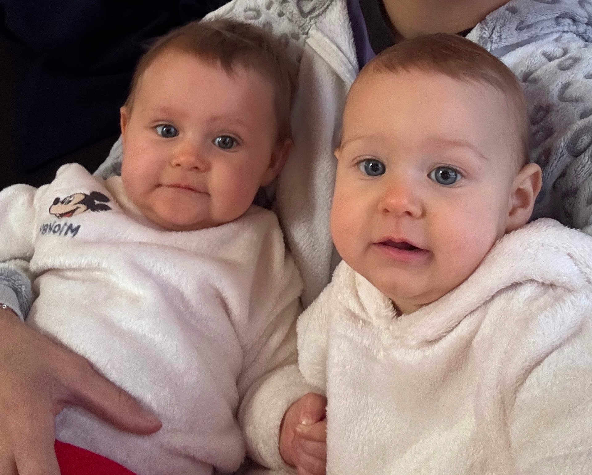 Lyla Et Maya a rejoint le concours — aidez-le/la à gagner de superbes lots ! baby, two_babies, infant, face, blue_eyes, chubby_cheeks, cozy_blanket, hoodie, white_clothing, mickey_mouse, holding_hands, sitting, indoor, couch, family_member, close_up, portrait, soft_texture, hand, adorable