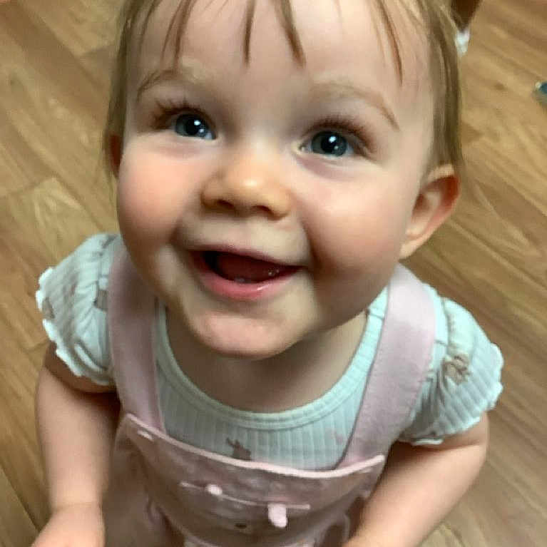 Alexis is registered to the contest to win money with this photo: toddler, child, smiling, happy, face, eyes, pink_clothing, overalls, indoor, wooden_floor, cute, portrait, baby, person, hair, looking_up, small_hands, cheerful, apparel, one_child