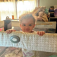 Amelia is registered to the contest to win money with this photo: toddler, child, playpen, blanket, curtains, window, indoor, smile, face, hands, mesh, fabric, hair, room, pile_of_clothes, baby, daylight, furniture, person, home