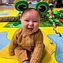 baby, child, smiling, frog_headband, knitted_clothing, indoor, playmat, colorful, happy, sitting, tongue_out, adult, toy, background, people, floor, cozy, cute, portrait, fun