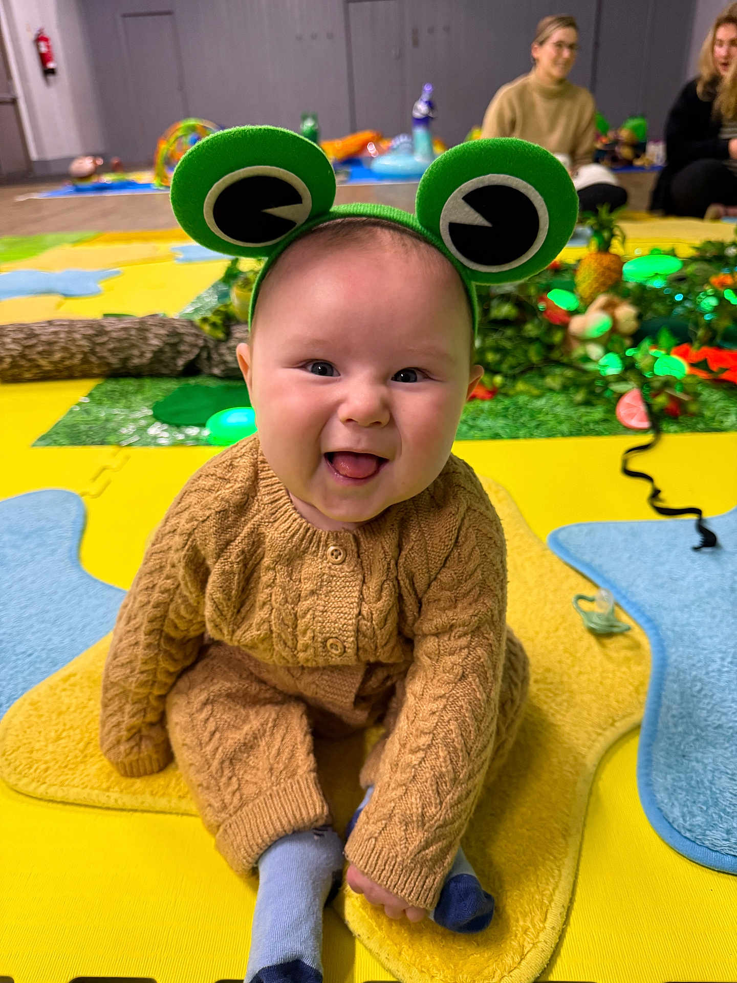 Alistair joined the competition — help win amazing prizes! baby, child, smiling, frog_headband, knitted_clothing, indoor, playmat, colorful, happy, sitting, tongue_out, adult, toy, background, people, floor, cozy, cute, portrait, fun