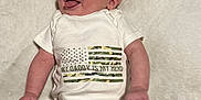 Creed is registered to the contest to win money with this photo: baby, infant, onesie, socks, camouflage, flag, tongue_out, smiling, lying_down, white_blanket, cute, playful, child, portrait, happy, soft_texture, clothing, person, indoor, newborn