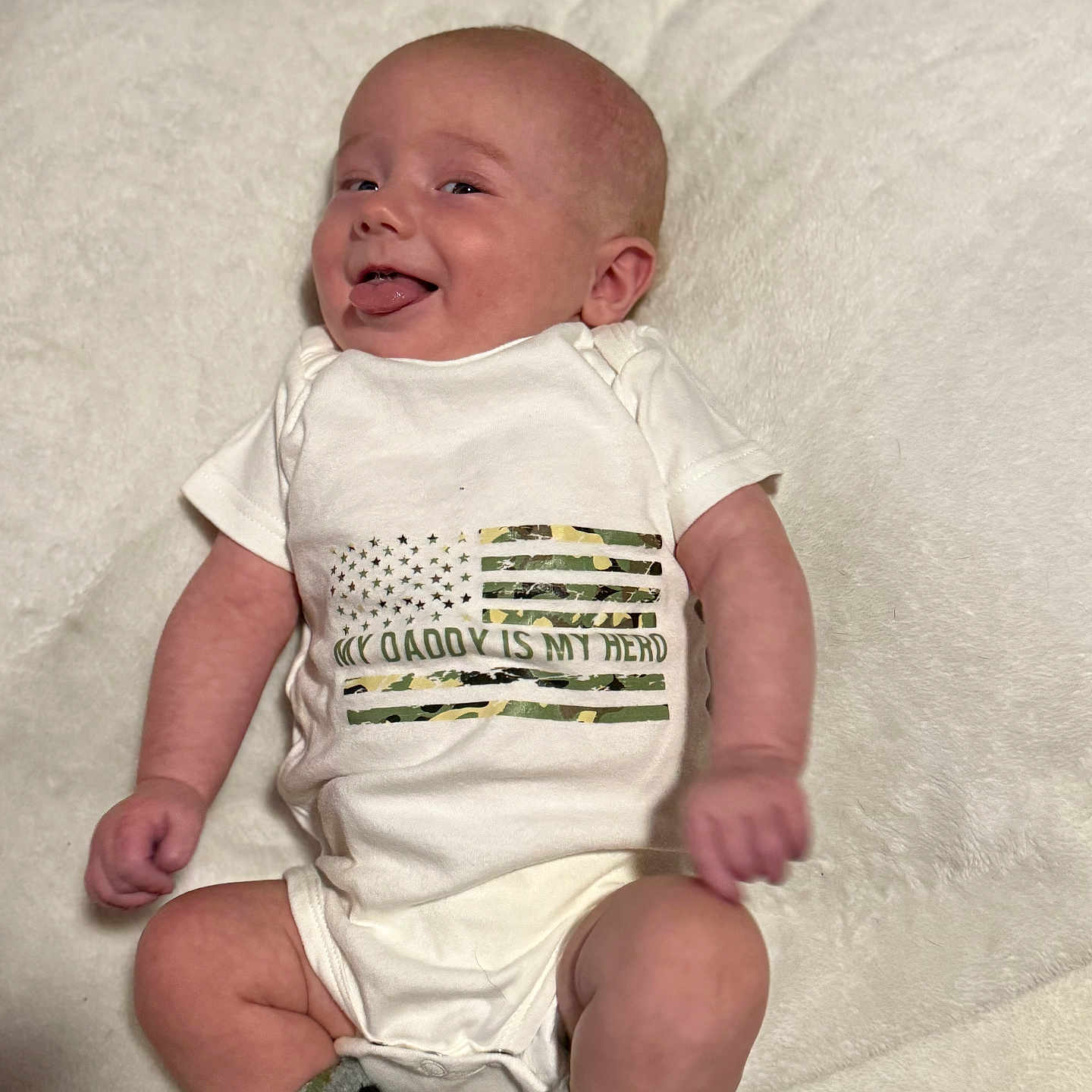 Creed is registered to the contest to win money with this photo: baby, camouflage, child, clothing, cute, flag, happy, indoor, infant, lying_down, newborn, onesie, person, playful, portrait, smiling, socks, soft_texture, tongue_out, white_blanket