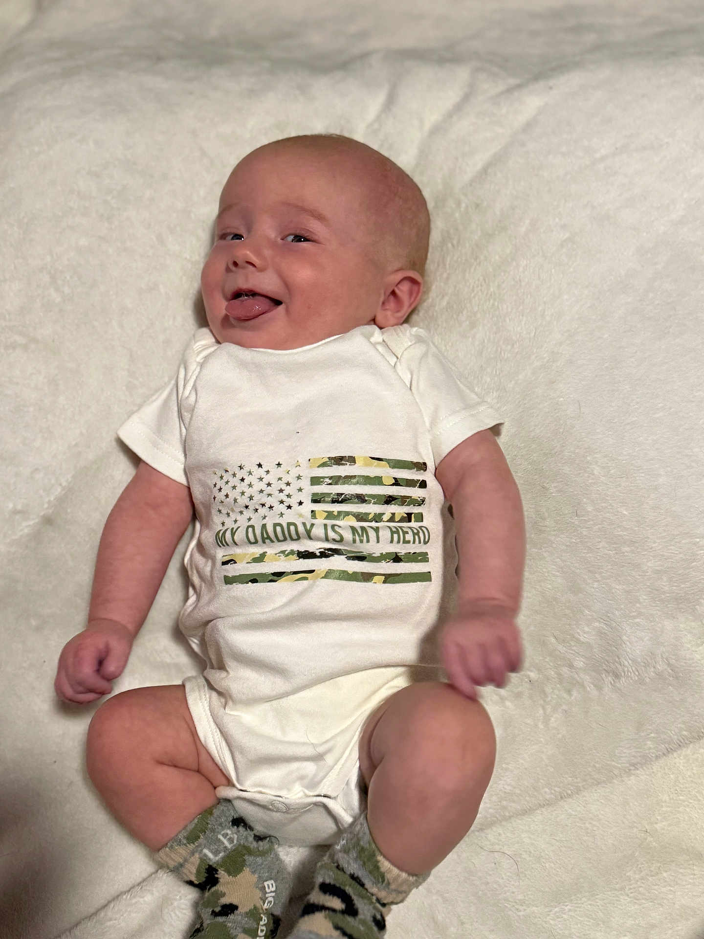Creed is registered to the contest to win money with this photo: baby, infant, onesie, socks, camouflage, flag, tongue_out, smiling, lying_down, white_blanket, cute, playful, child, portrait, happy, soft_texture, clothing, person, indoor, newborn