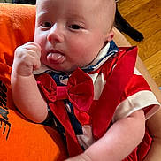 Creed is registered to the contest to win money with this photo: baby, infant, face, tongue, red_bow_tie, clothing, hand, person, orange_shirt, dog, wooden_floor, indoor, cute, playful, closeup, portrait, human, sitting, holding, expression
