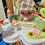 baby, child, pacifier, hat, plaid, green_shirt, activity_center, toy, button, hand, face, eyes, indoor, colorful, play, infant, seat, pattern, fabric, curious