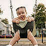 baby, child, swing, playground, outdoor, smiling, tongue_out, happy, legs, sandals, trees, houses, sidewalk, chains, clothing, green_blanket, sunset_light, face, person, fun